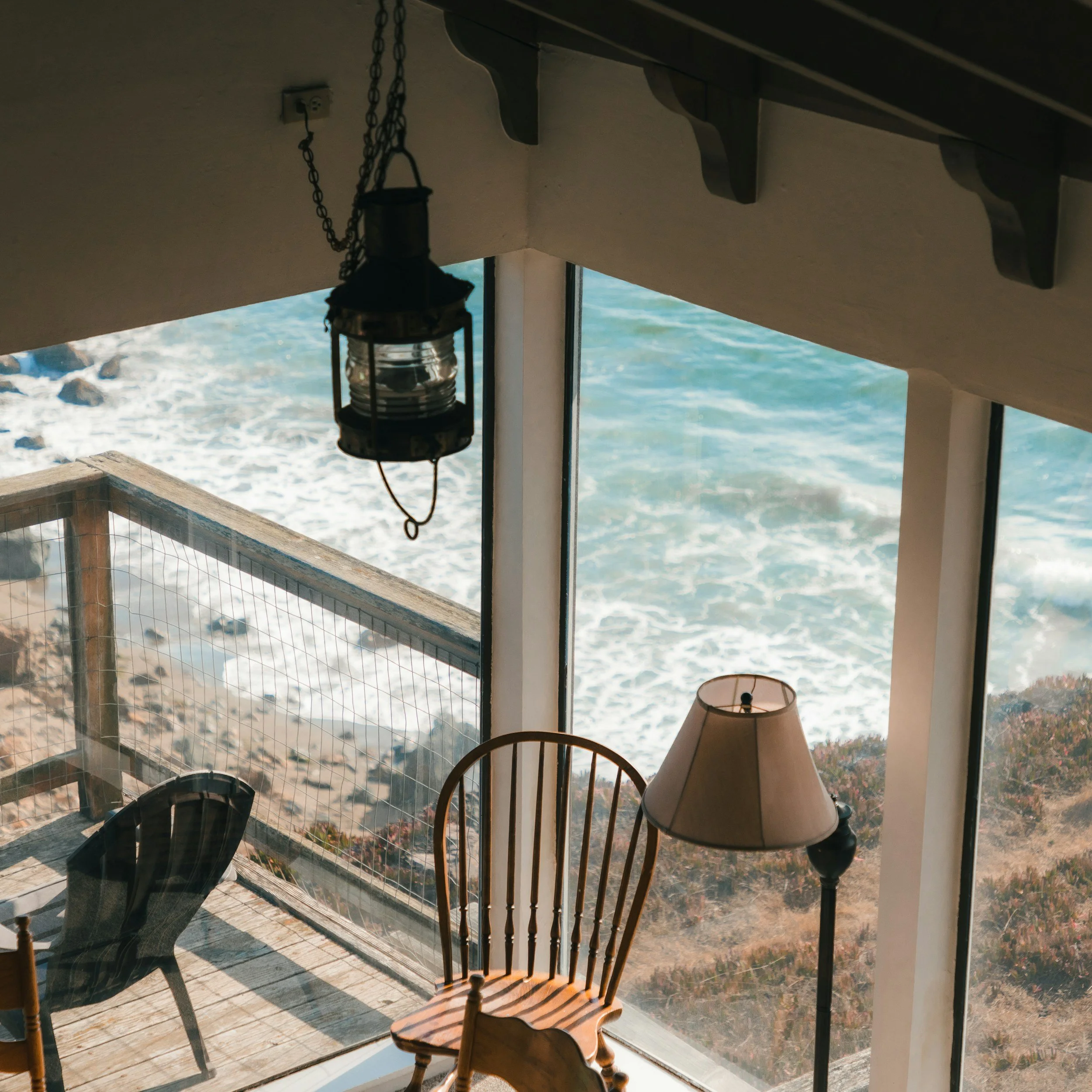 A wooden indoor staircase near a large window overlooking a shoreline with water and rocks. There is a hanging lantern near the window, a lamp on a table, and a chair in the foreground.
