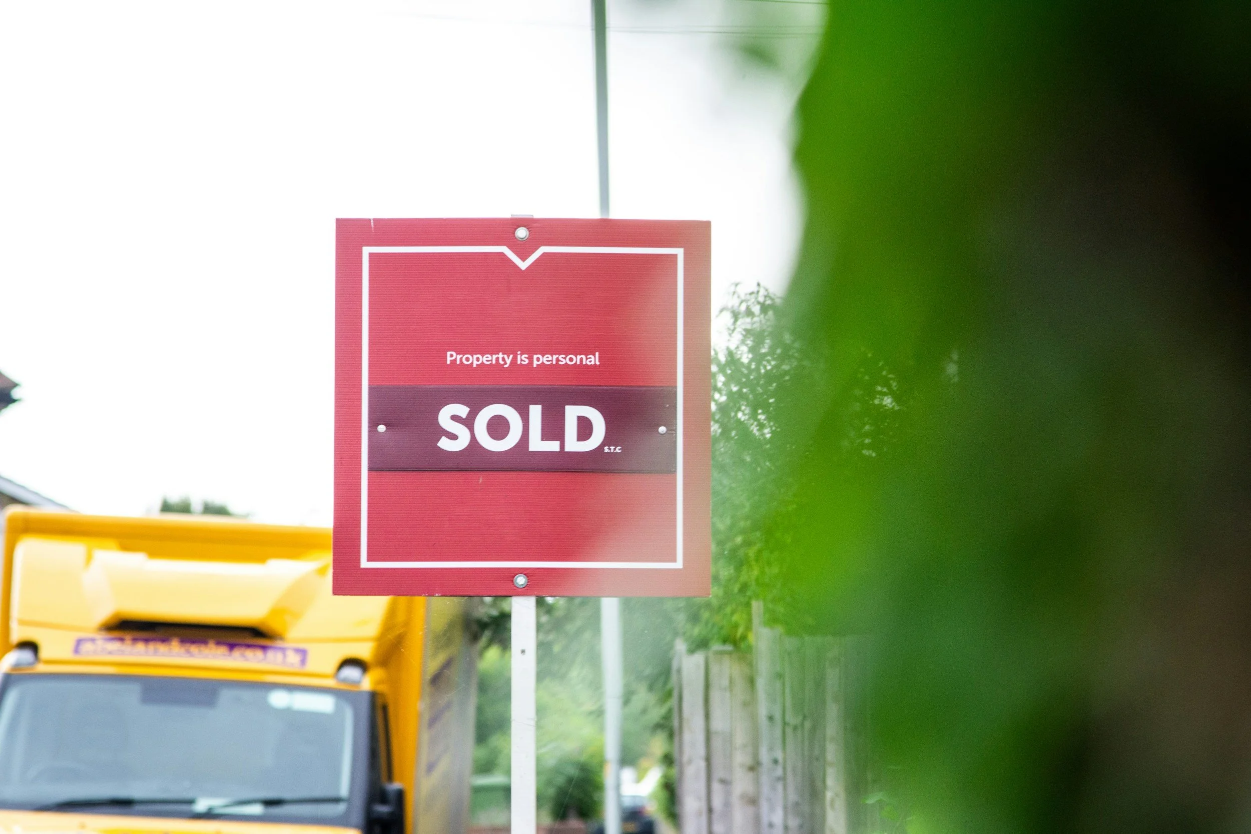 A red property for sale sign with white text reading "Property is personal" and "SOLD" underneath, situated outdoors with a green plant partially obscuring the view on the right and a yellow vehicle in the foreground.