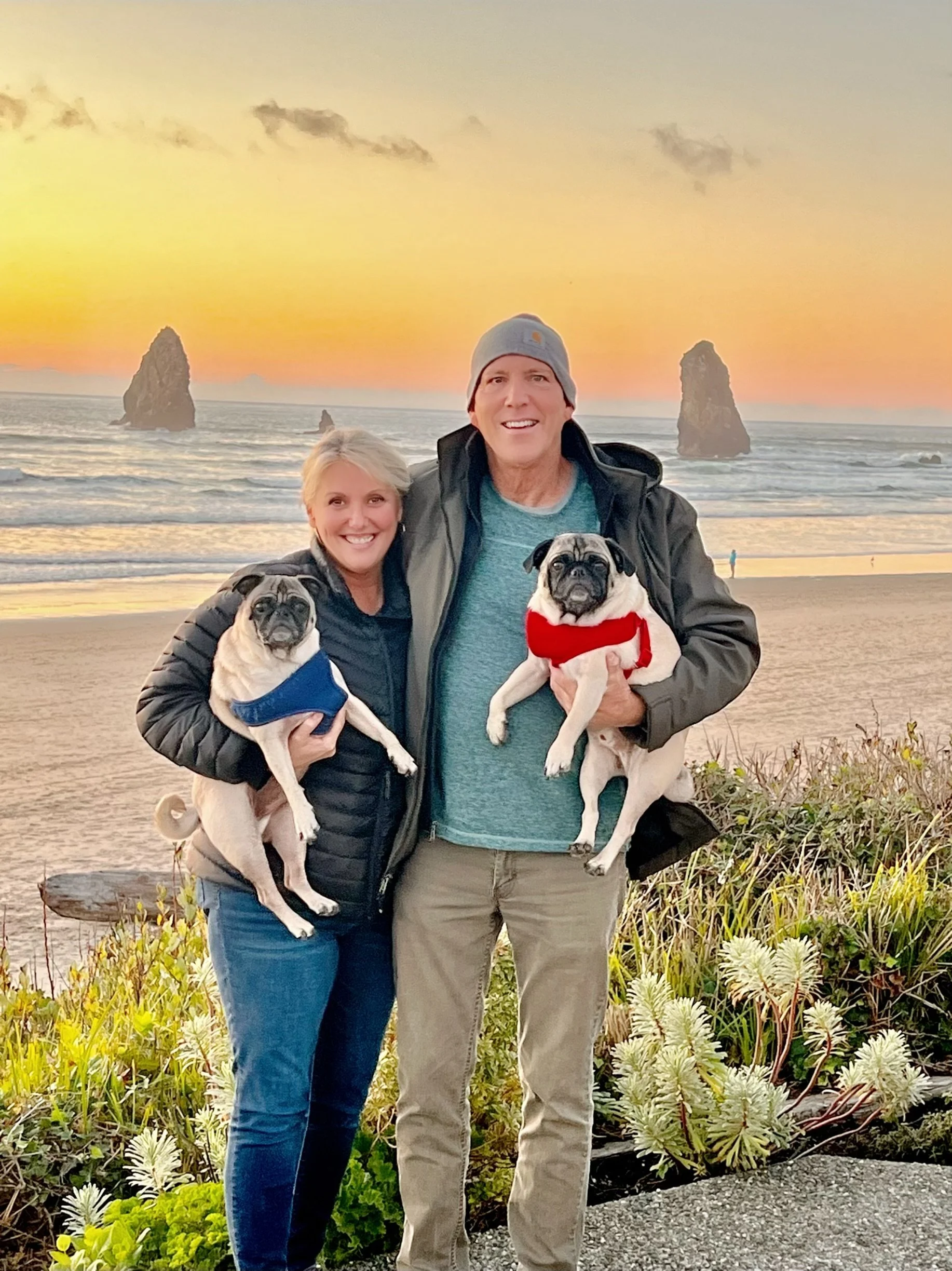 Beth Flora, Oregon Coast realtor, in Cannon Beach, holding a pug, with man is wearing a gray beanie and gray jacket. In the background, haystack Rock, and Needles rocks in the ocean and a colorful sunset sky.