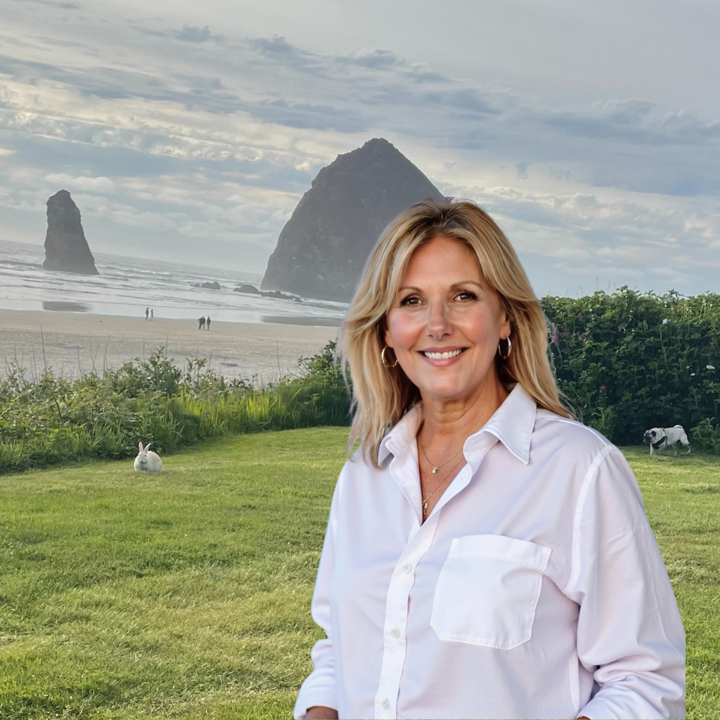 Beth Flora, Oregon Coast real estate agent serving Cannon Beach and Seaside with blonde hair smiling at the camera, wearing a white shirt, standing on the beach in Cannon Beach, with Haystack Rock with a rabbit and a dog on the grassy area.