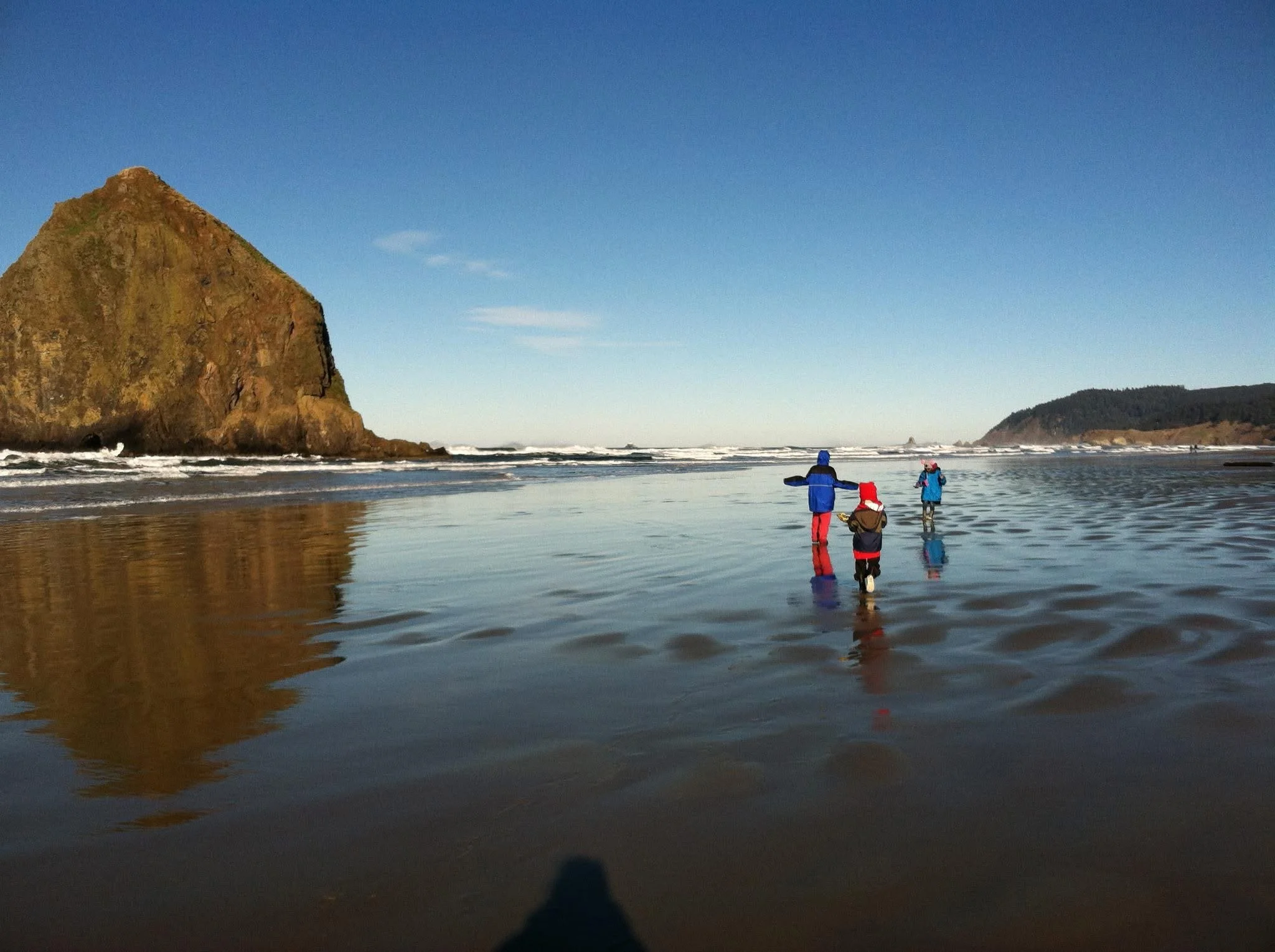 Three children in colorful jackets and hats walking on a wet sandy beach Cannon Beach Oregon, Haystack Rock large rock formation in the background.