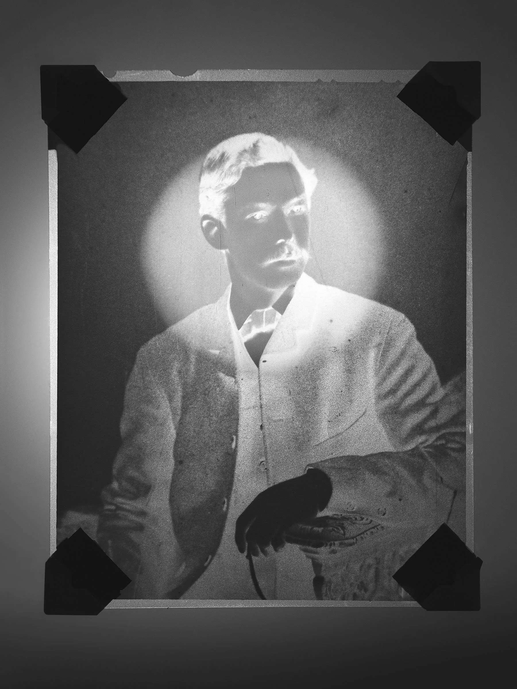 Black and white projection of a man with light hair, wearing a button-up shirt, resting his hand on a surface, with the image taped at the corners.