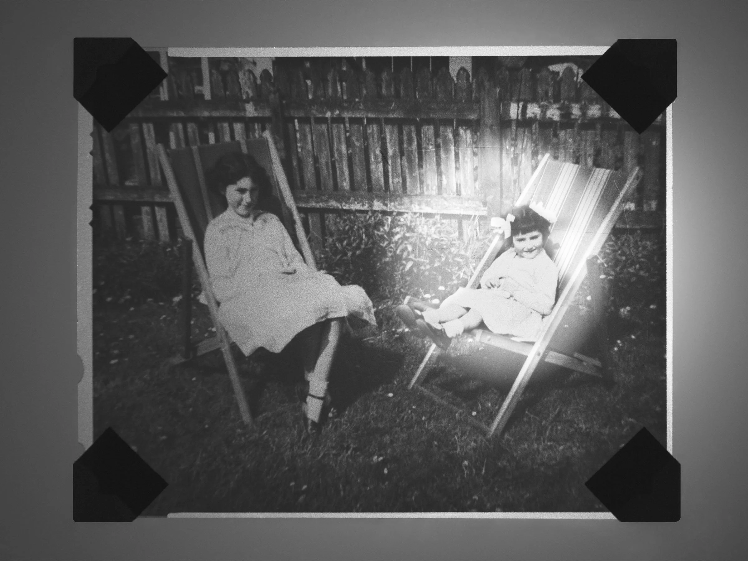 Black and white photo of a woman and a young girl sitting on outdoor chairs in a backyard with a wooden fence in the background.