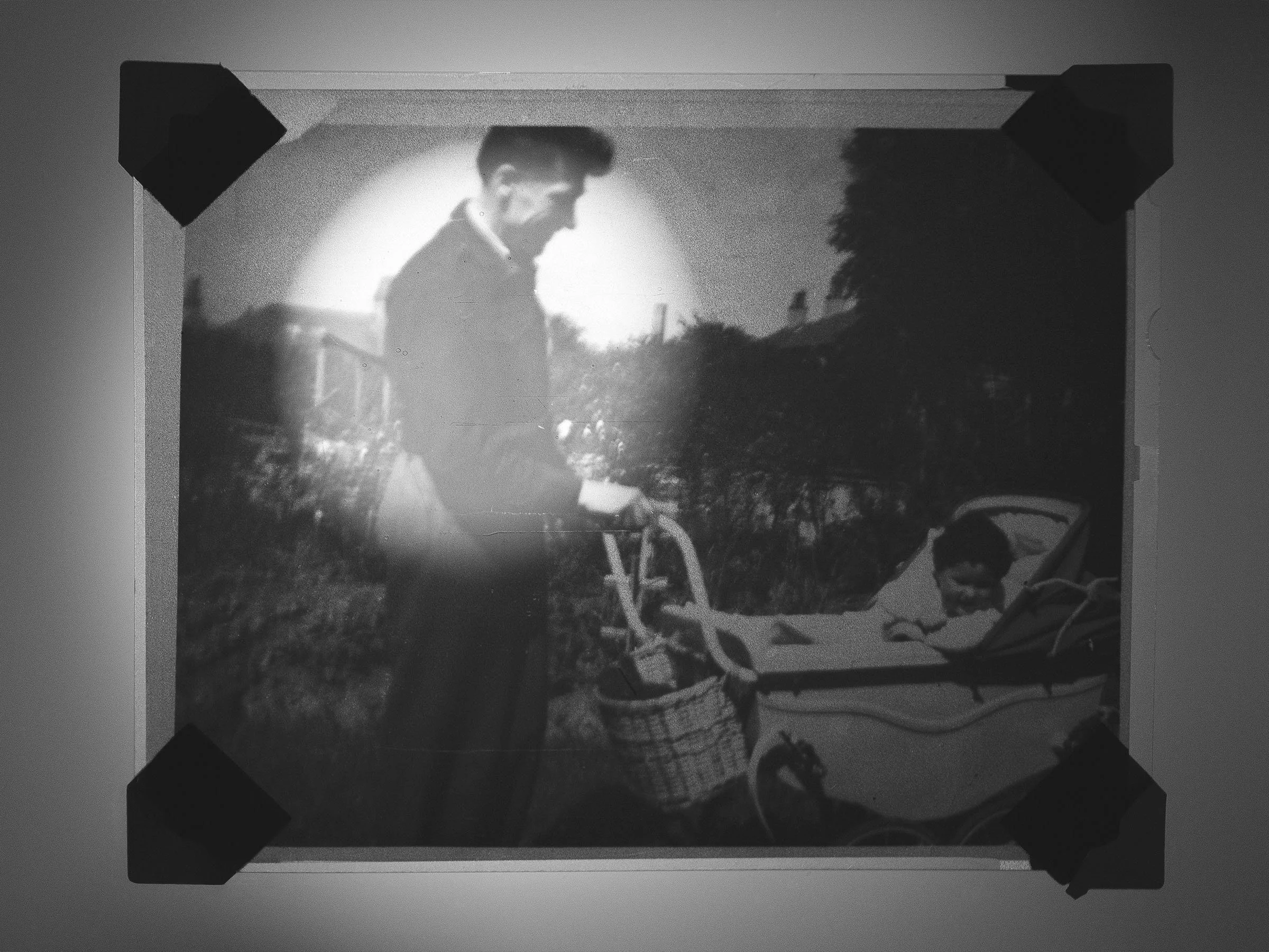 A young man pushing a stroller with a child lying inside, outdoors during daytime, with trees and houses in the background.