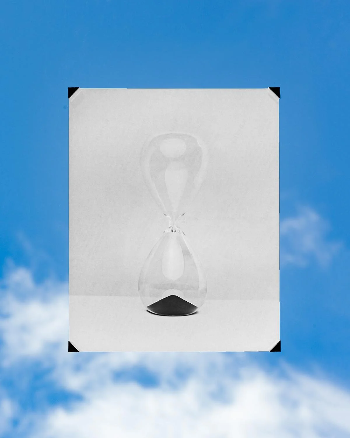 An hourglass with black sand on the bottom, seen through a transparent glass, against a blue sky with scattered clouds.