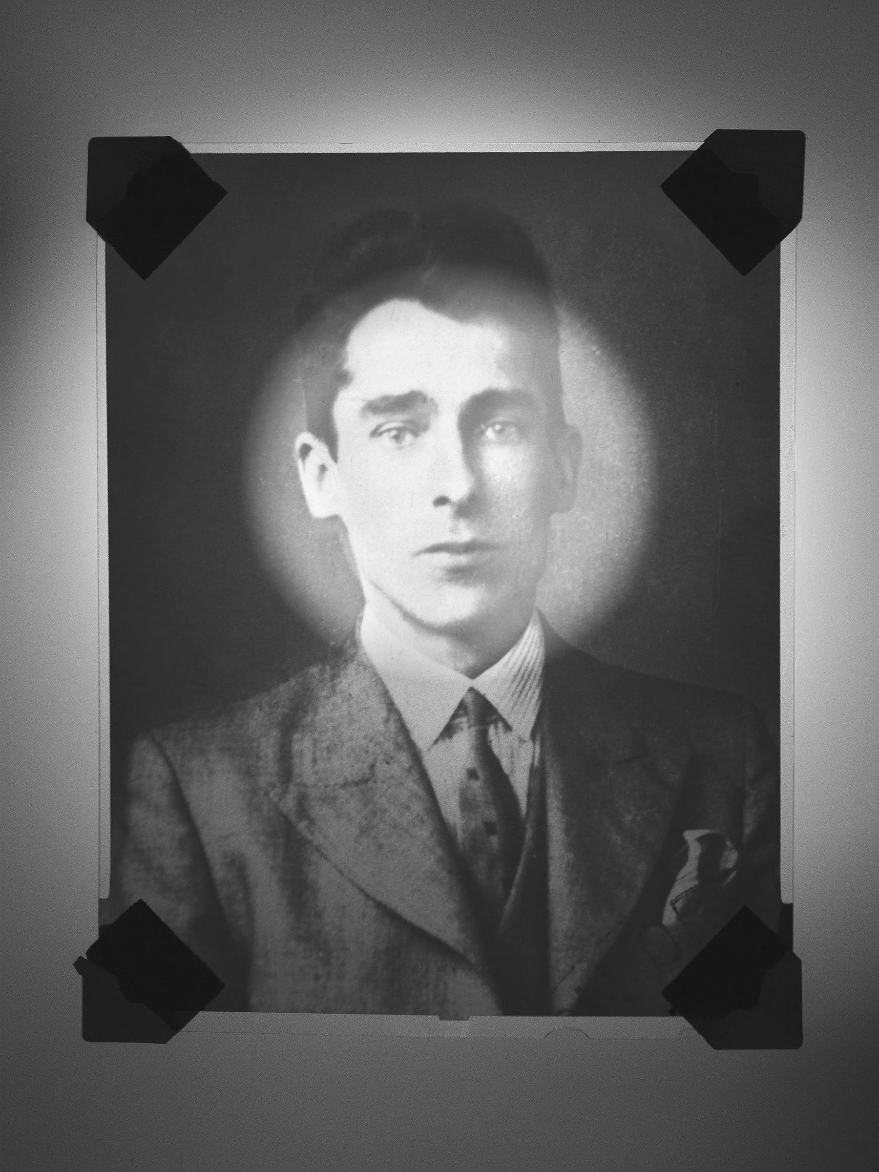 A black-and-white portrait photograph of a young man in formal attire, pinned with black tape on each corner.