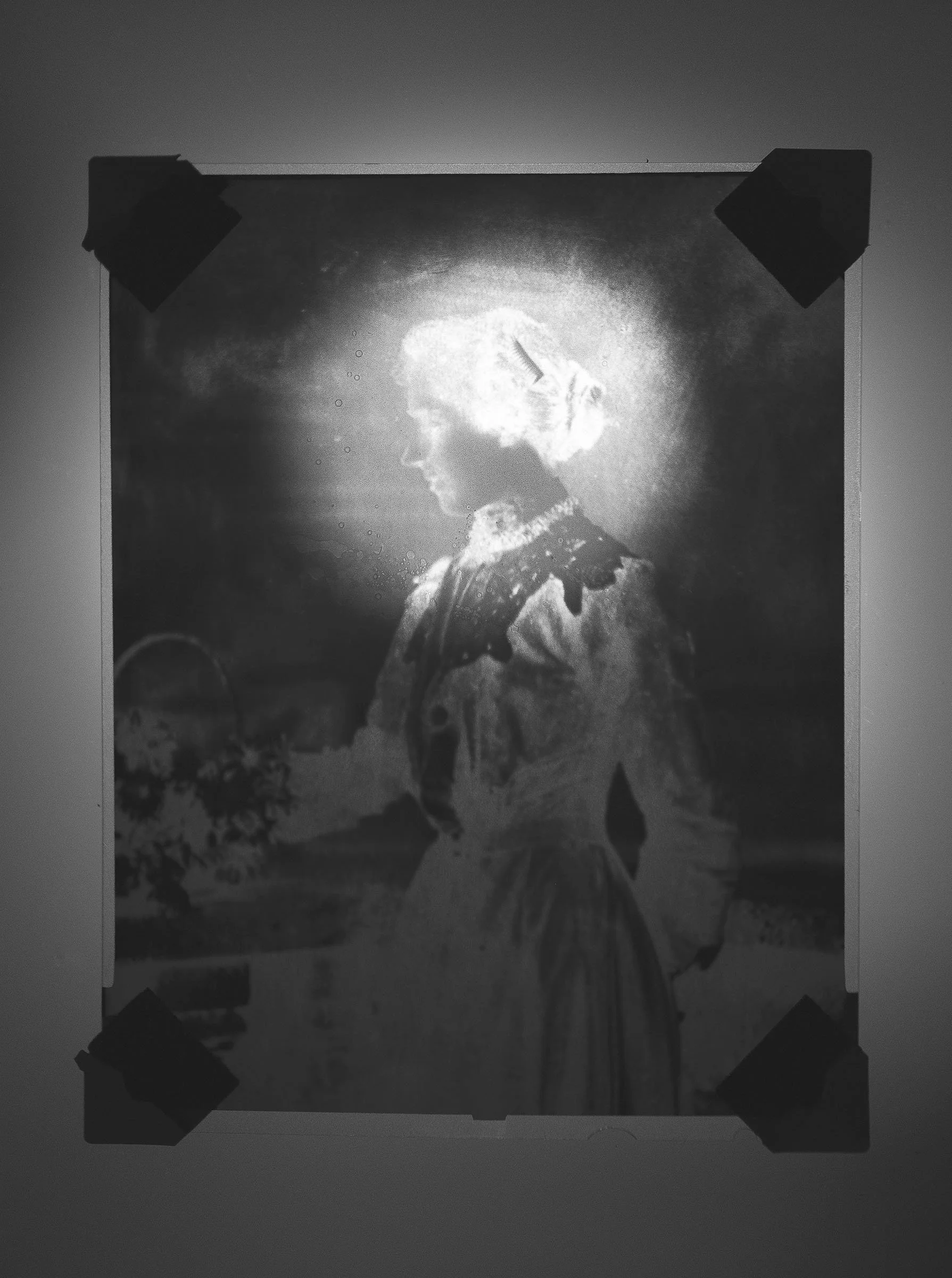 Black-and-white photograph of a woman in vintage clothing, facing left, with her hair in an updo. The background is dark with a bright light source illuminating her profile from behind.