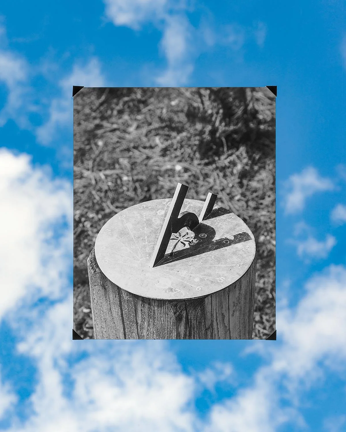 Black and white photograph of a clock face with three-dimensional numbers 4, 5, and 6, mounted on a wooden post, with a background of dirt and grass.