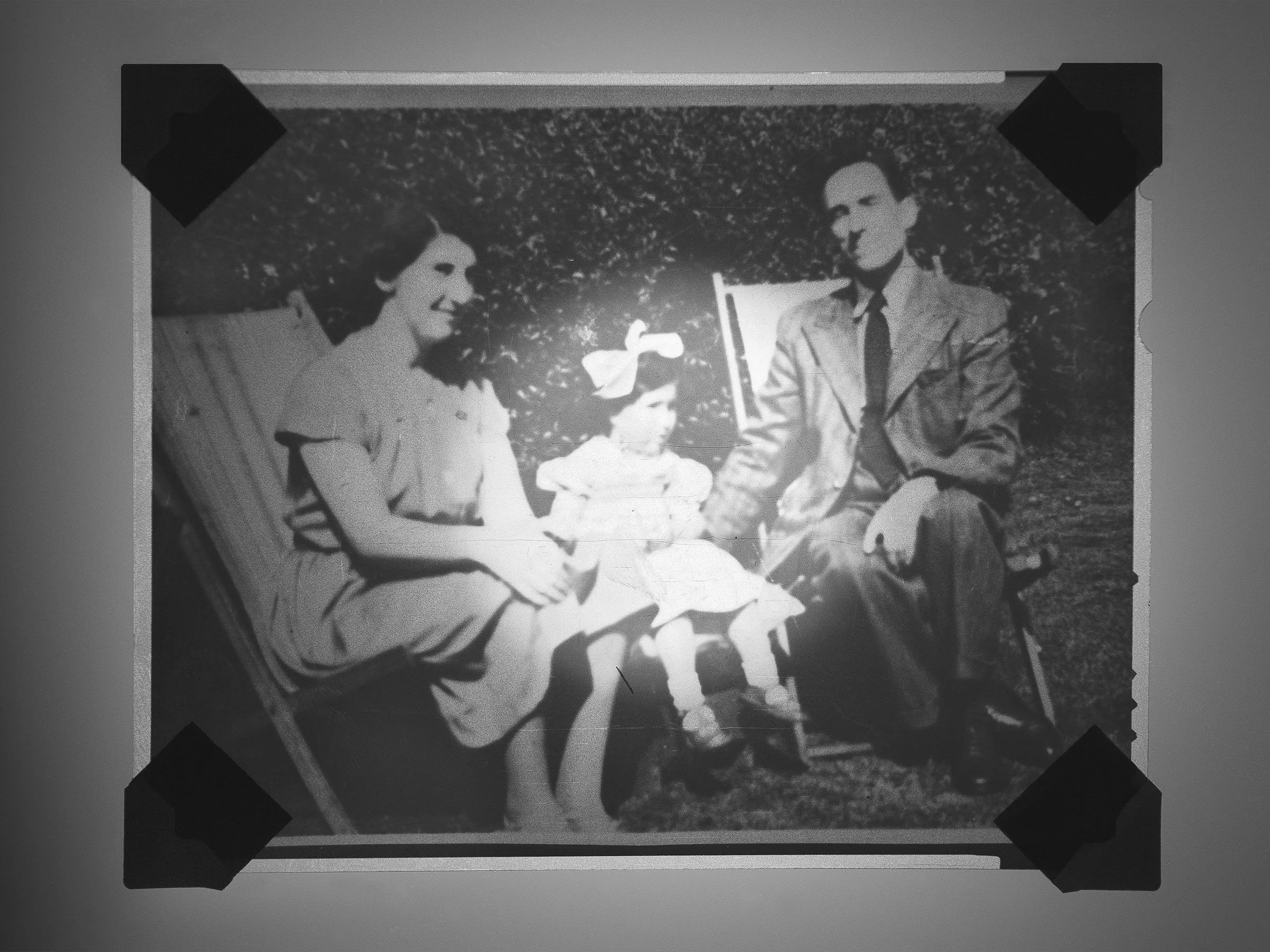Black and white photo of a woman, young girl, and man sitting outdoors with a dog in the center, in an old-fashioned setting.