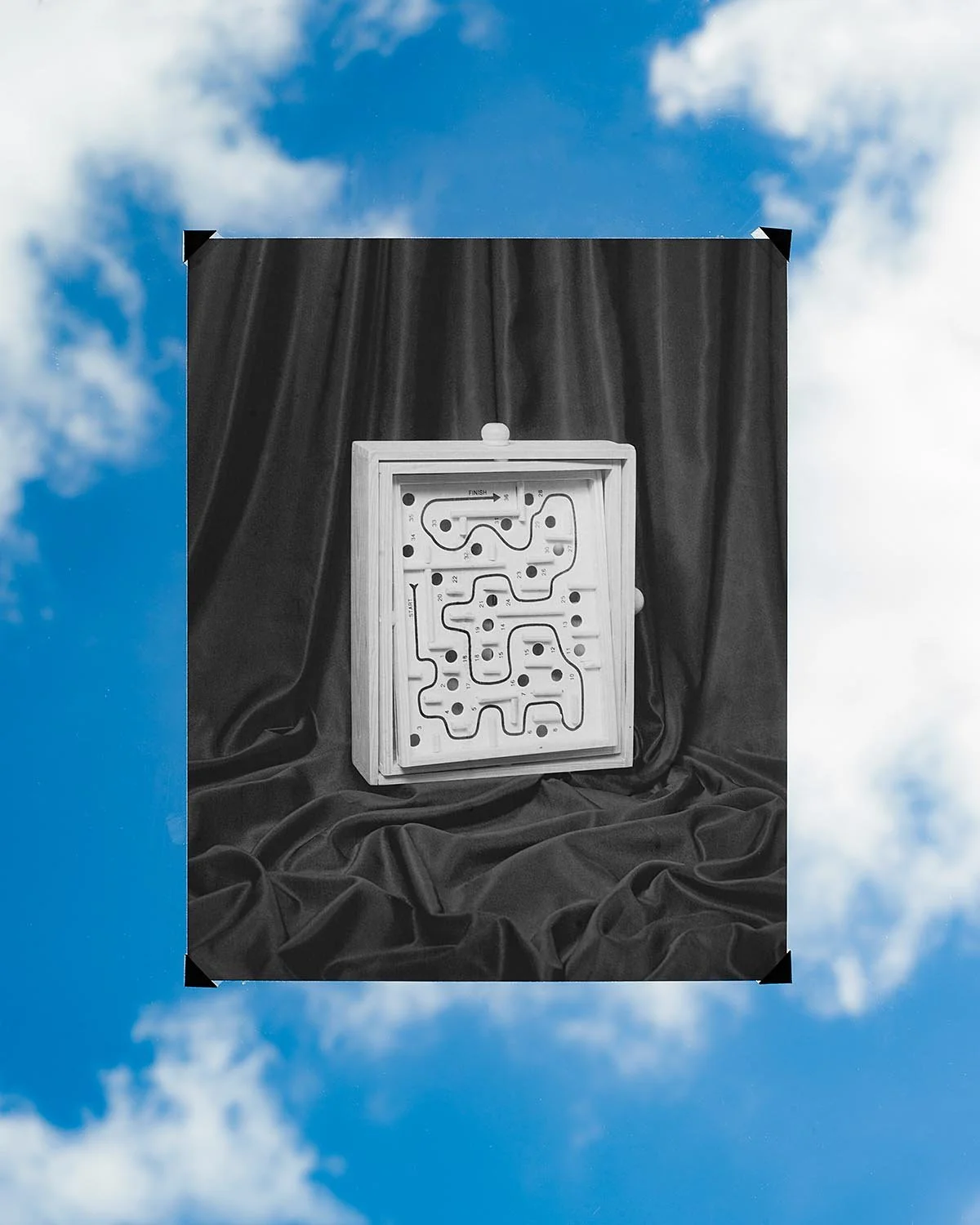 A maze game board with pathways and holes, placed on black fabric with cloud sky background.