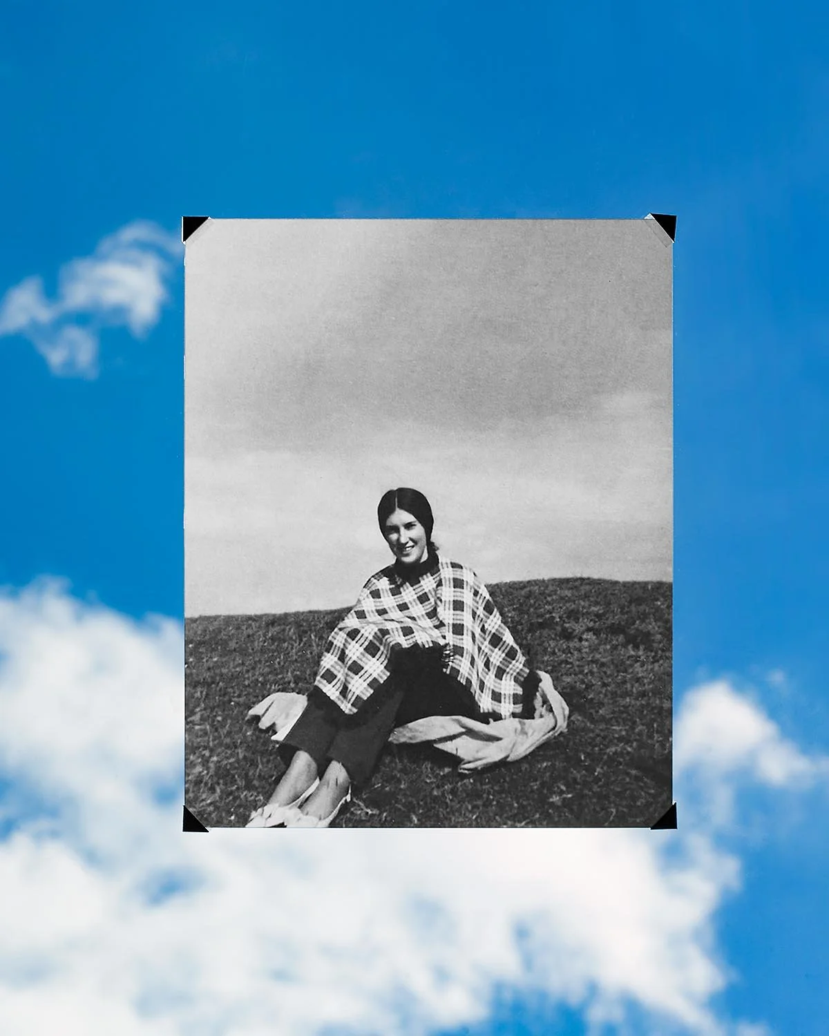 Black and white photograph of a woman sitting on grass, smiling, with a landscape of hills behind her.