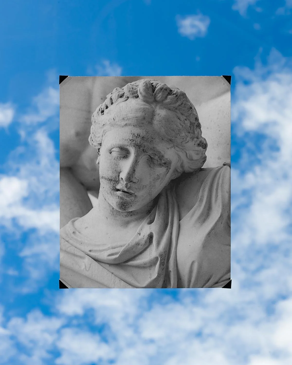 Black and white photograph of a classical marble sculpture of a woman's head and upper body, with a sheathed garment and flowing hair, against a cloudy sky background.