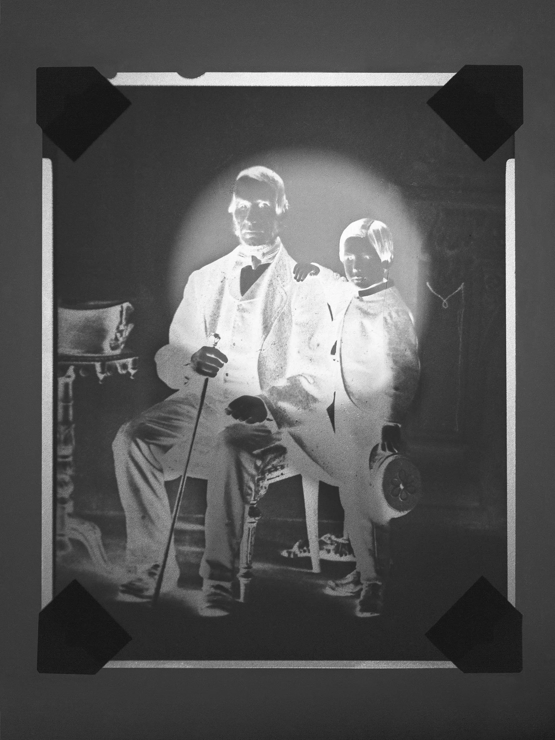 Black and white photograph of a man sitting with one leg crossed over the other and a young boy standing beside him, both dressed in vintage clothing, with their arms around each other's shoulders.