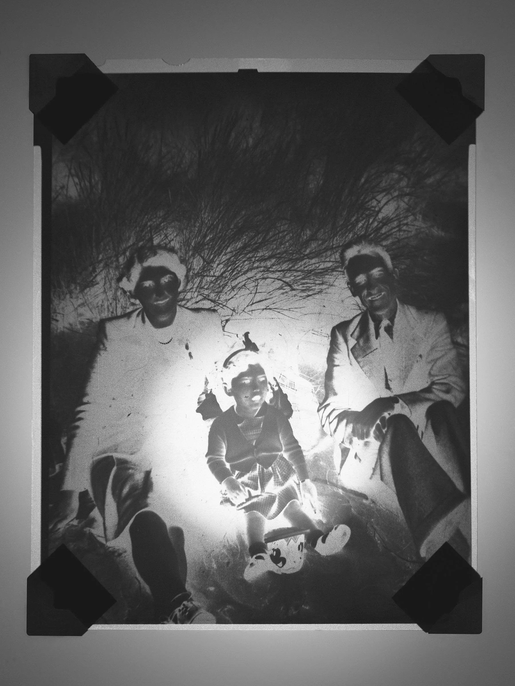 Black and white photo of a family of three outdoors, with two adults and one child. They are sitting or lying on the ground, with trees and branches in the background.