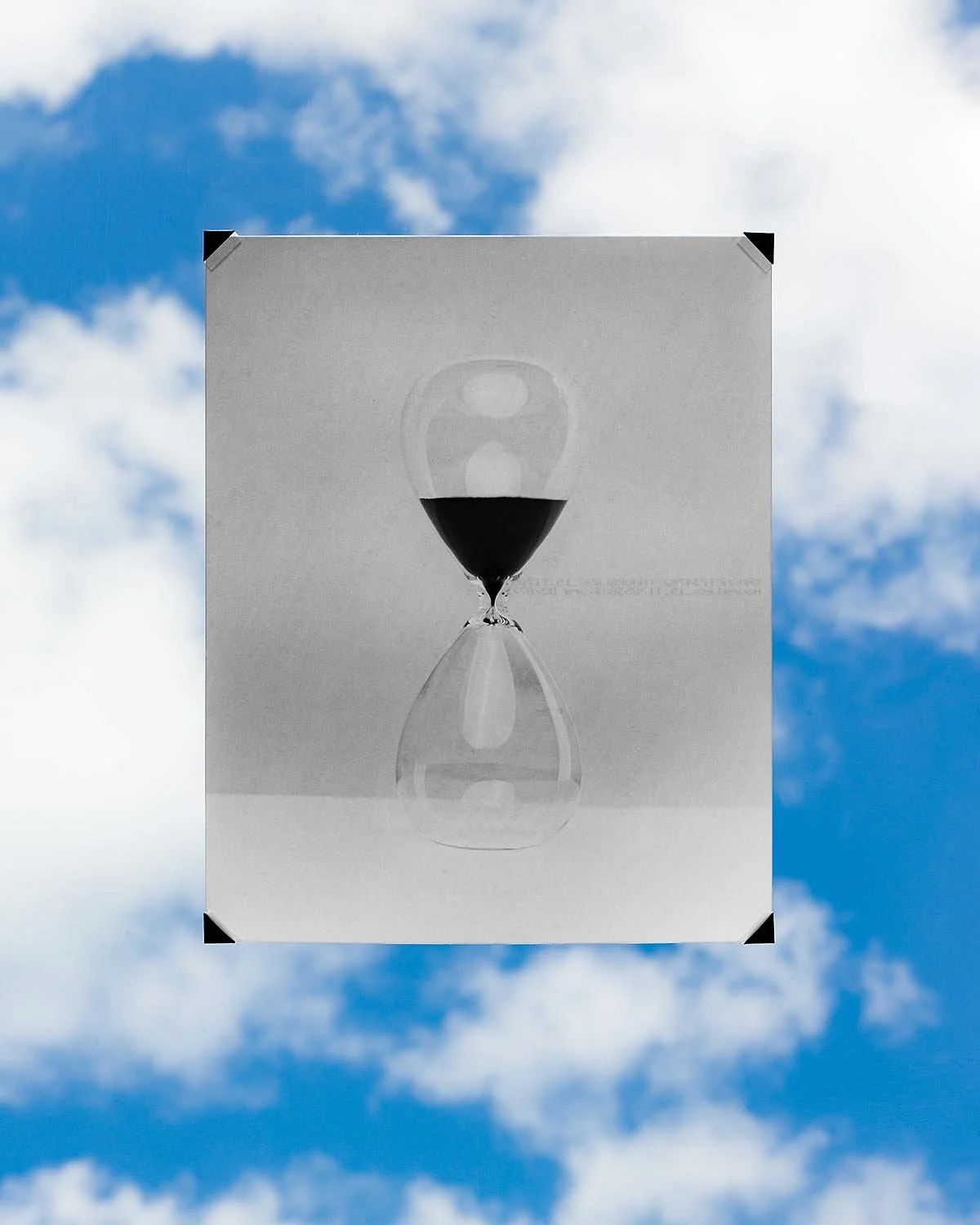A black and white photograph of an hourglass with sand in the upper chamber, reflected upside down on a reflective surface, with a cloudy blue sky in the background.