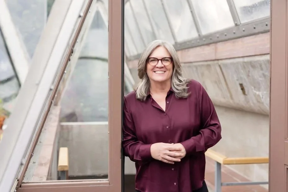 A smiling woman with gray hair and glasses standing inside a greenhouse, leaning against a doorframe, wearing a burgundy blouse.