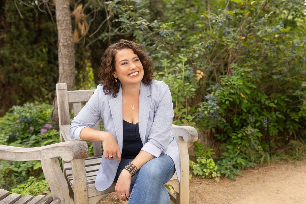 A woman with curly brown hair, wearing a light gray blazer over a black top and jeans, sitting on a wooden park bench outdoors with green trees and bushy plants in the background, smiling.