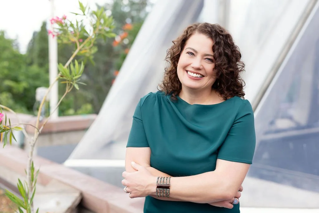 A woman with curly brown hair wearing a teal dress, smiling outdoors on a balcony with plants and a glass structure in the background.