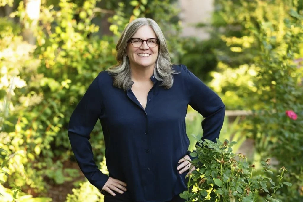 A woman with gray hair and glasses smiling, standing outdoors in a garden with green plants and bushes.
