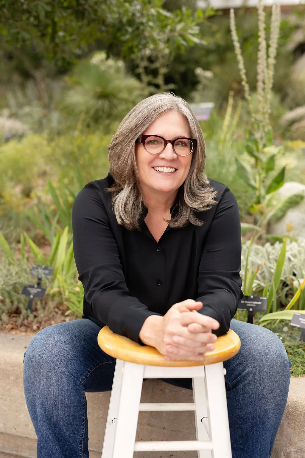 A woman with shoulder-length gray hair and glasses, wearing a black shirt, sitting on a white stool outdoors with greenery in the background, smiling at the camera.