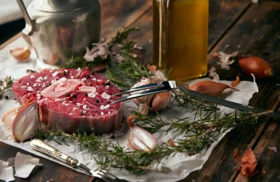 Raw meat on butcher paper with garlic cloves, herbs, a bottle of olive oil, and kitchen utensils on a wooden table.
