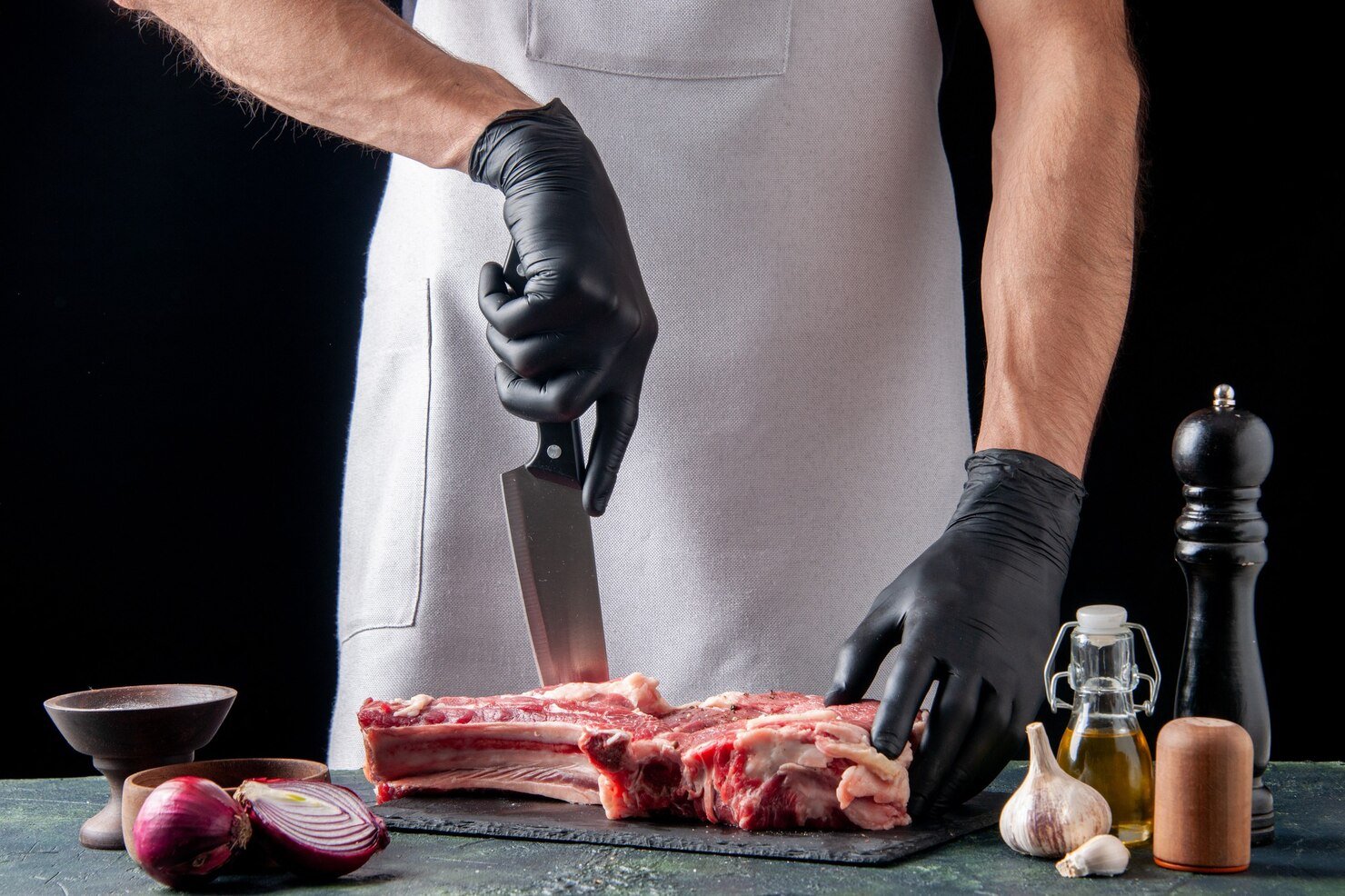 Person wearing black gloves and a gray apron slicing a large piece of raw meat on a black cutting board, surrounded by garlic, onion, oil, pepper grinder, and salt shaker, on a dark green surface against a black background.