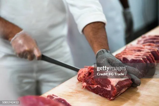 Person slicing raw meat on a wooden cutting board with a knife, wearing gloves.