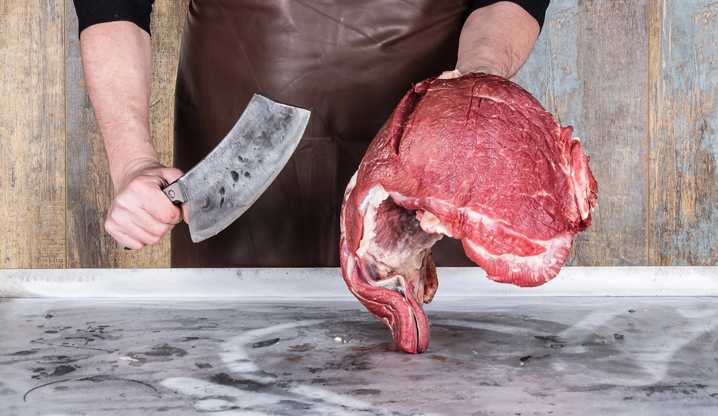 A person holding a cleaver next to a large piece of raw meat with a bone, on a butcher's table.