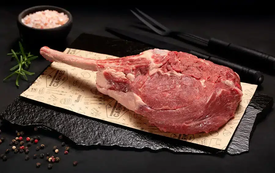 Raw beef tomahawk steak on black slate with peppercorns, salt, and black-handled cutlery on dark background.