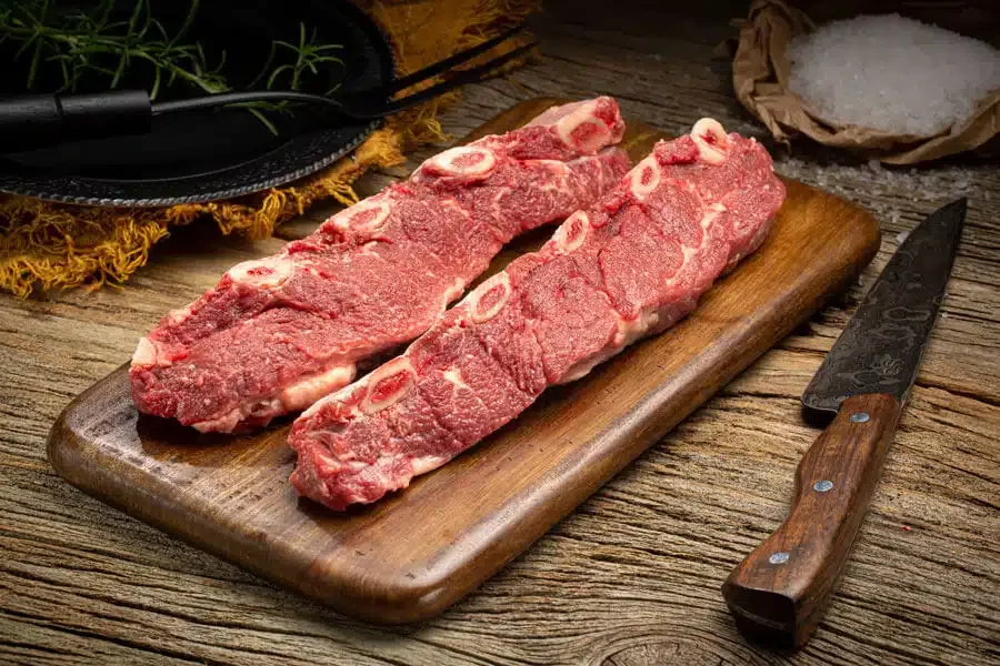 Two raw beef ribeye steaks on a wooden cutting board with a knife nearby, salt in a paper bag, and herbs in the background.