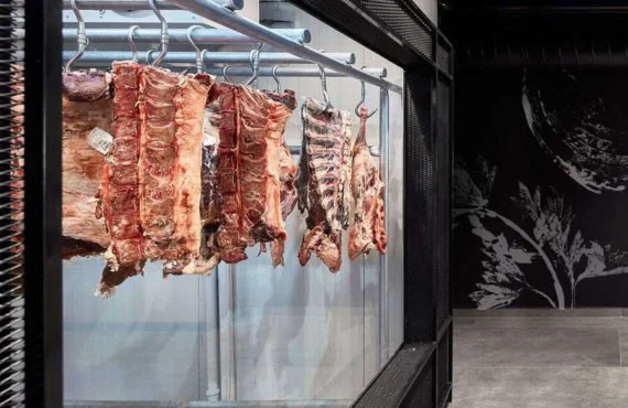 Various cuts of raw meat hanging inside a meat storage or display case at a butcher shop or meat market.