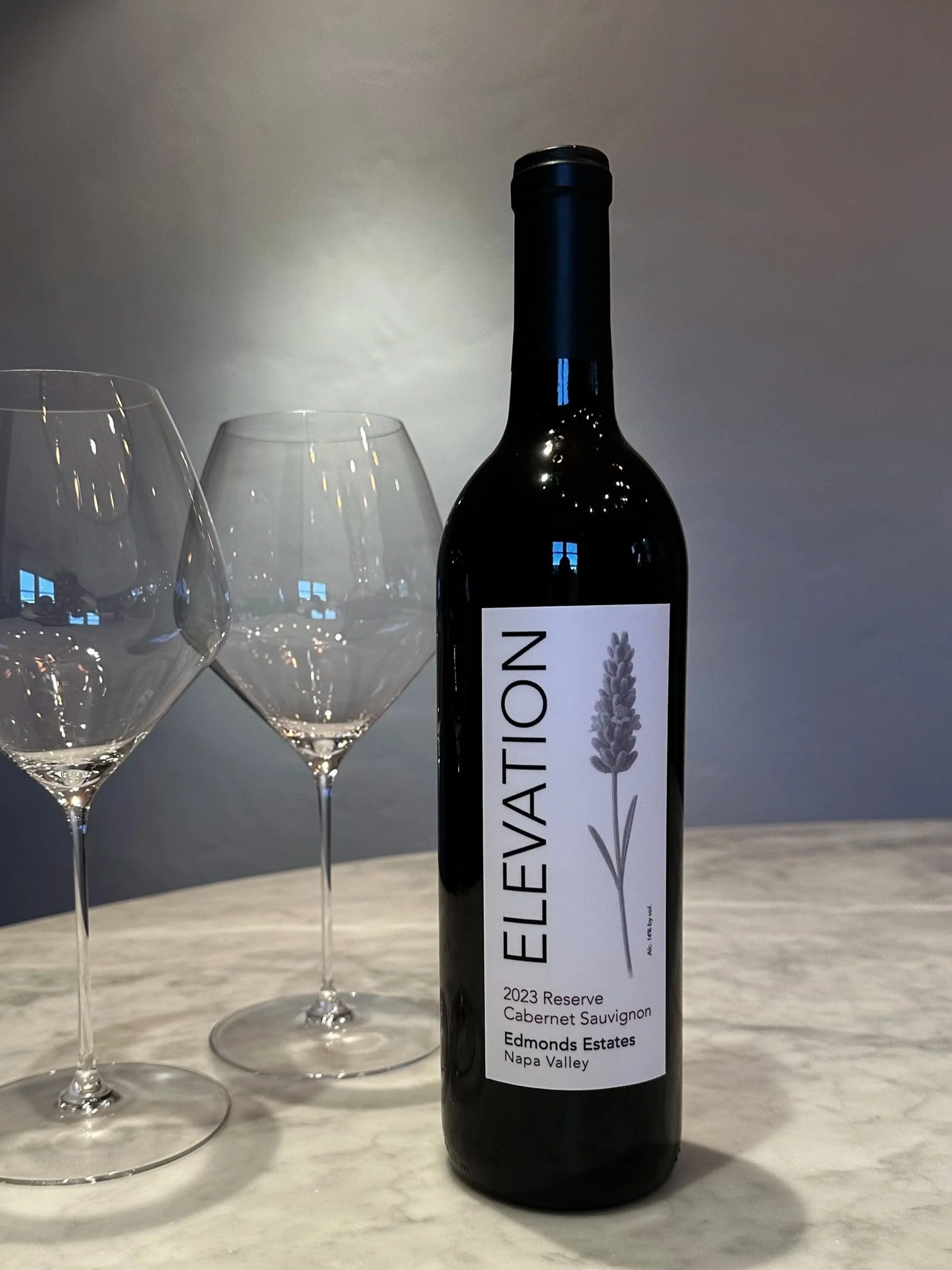 A bottle of Elevation 2023 Reserve Cabernet Sauvignon wine placed on a marble countertop next to two empty wine glasses.
