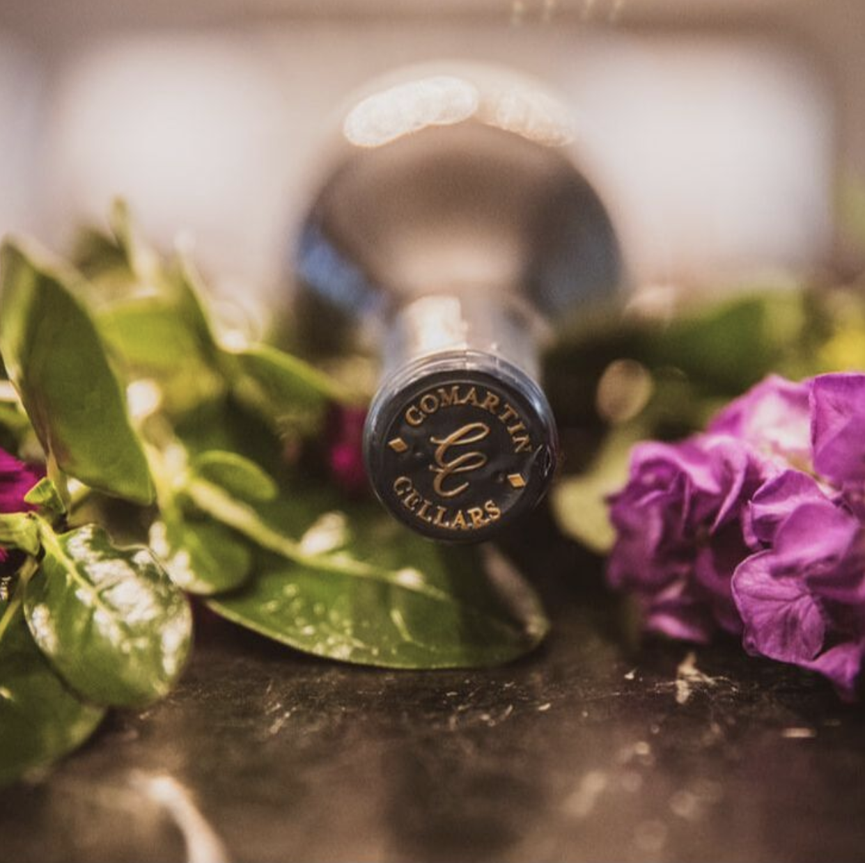 A bottle of Comartin Gillars wine lying on a surface surrounded by green leaves and purple flowers.