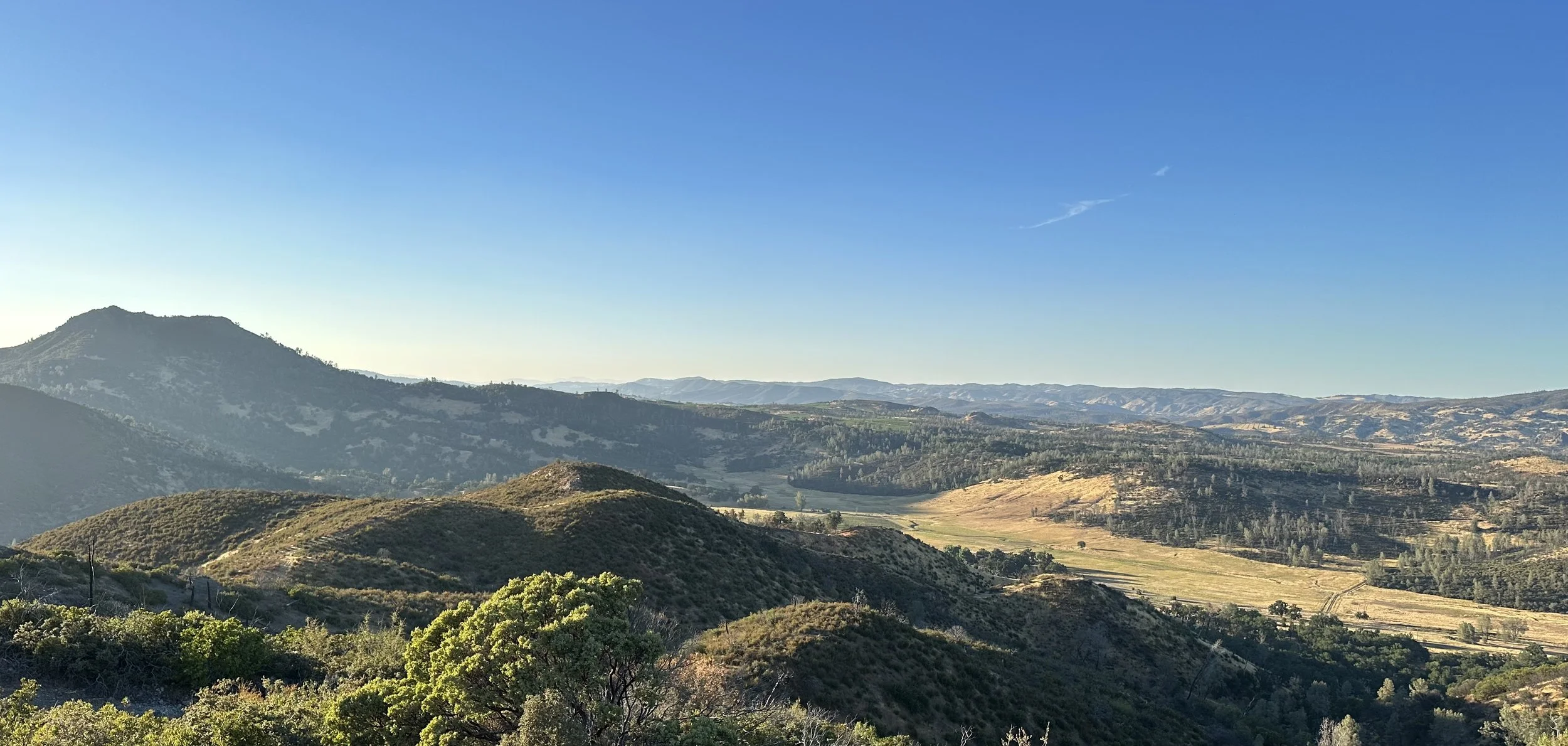 Scenic view of rolling hills and mountains under a clear blue sky with some thin clouds.