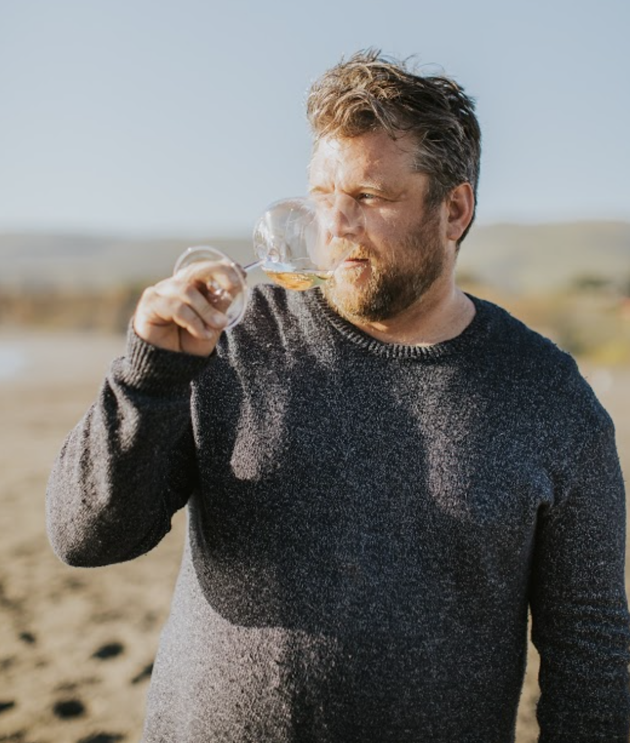 A man with a beard wearing a dark sweater, outdoors in a sunny field, inspecting a glass of amber-colored wine or whiskey.