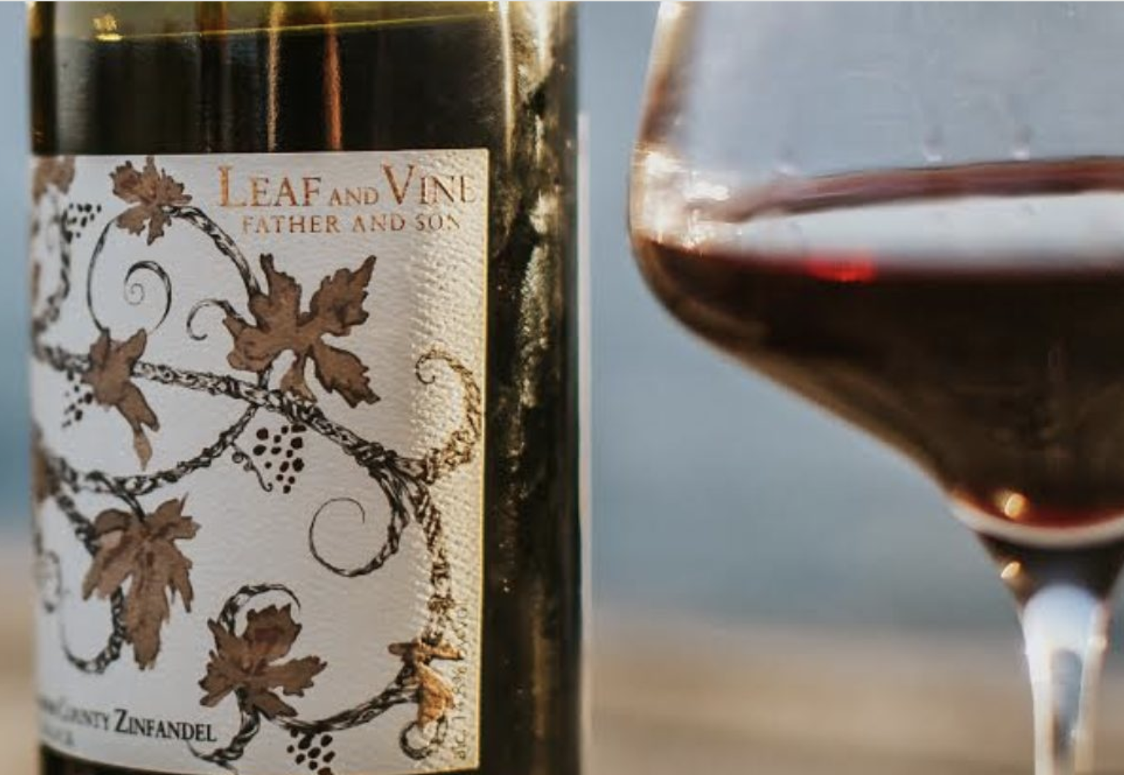 Close-up of a wine bottle labeled 'Leaf and Vine, Father and Son' with an illustration of grapevines, and a glass of red wine partially visible on the right.