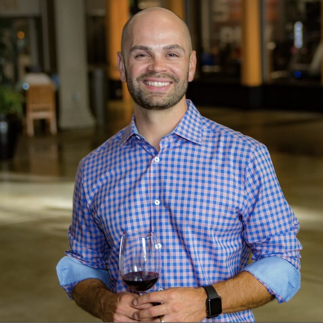 A man with a bald head and beard smiling while holding a glass of red wine, wearing a blue and pink checkered shirt with rolled-up sleeves, in an indoor setting.
