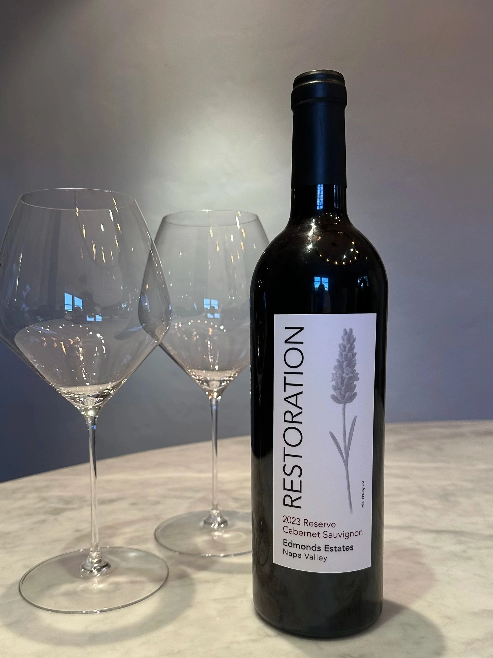 A bottle of Restoration 2023 Reserve Cabernet Sauvignon from Edmonds Estates, Napa Valley, positioned on a white surface with two empty wine glasses behind it.