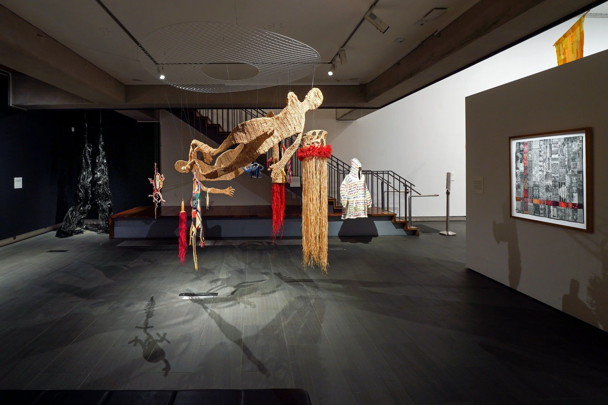 Art installation of various hanging sculptures, including a large dog figure made of woven material, a human figure, and abstract pieces in a gallery space.