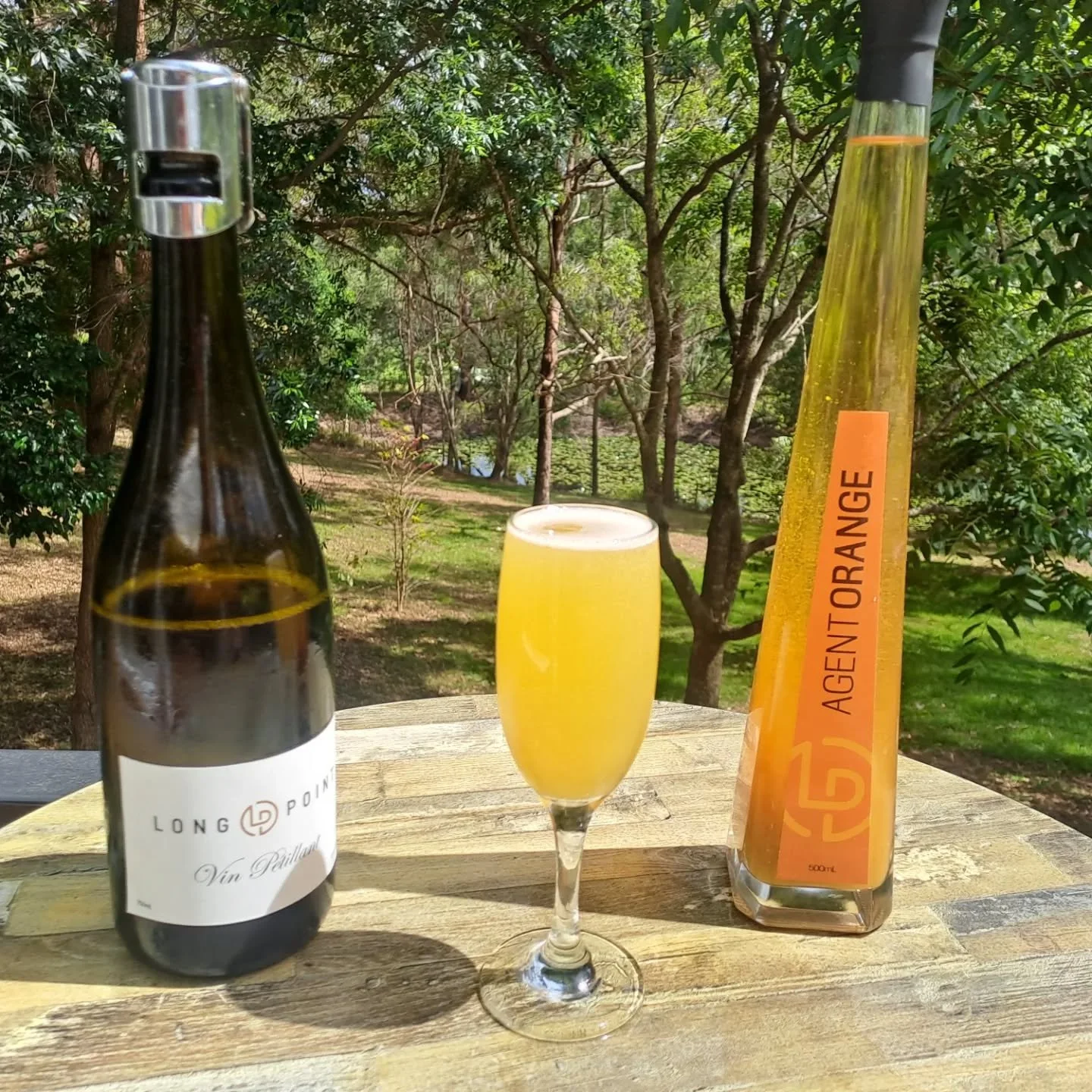 A bottle of wine, a glass of orange beverage, and a bottle of orange-flavored syrup on a wooden table outdoors with trees in the background.