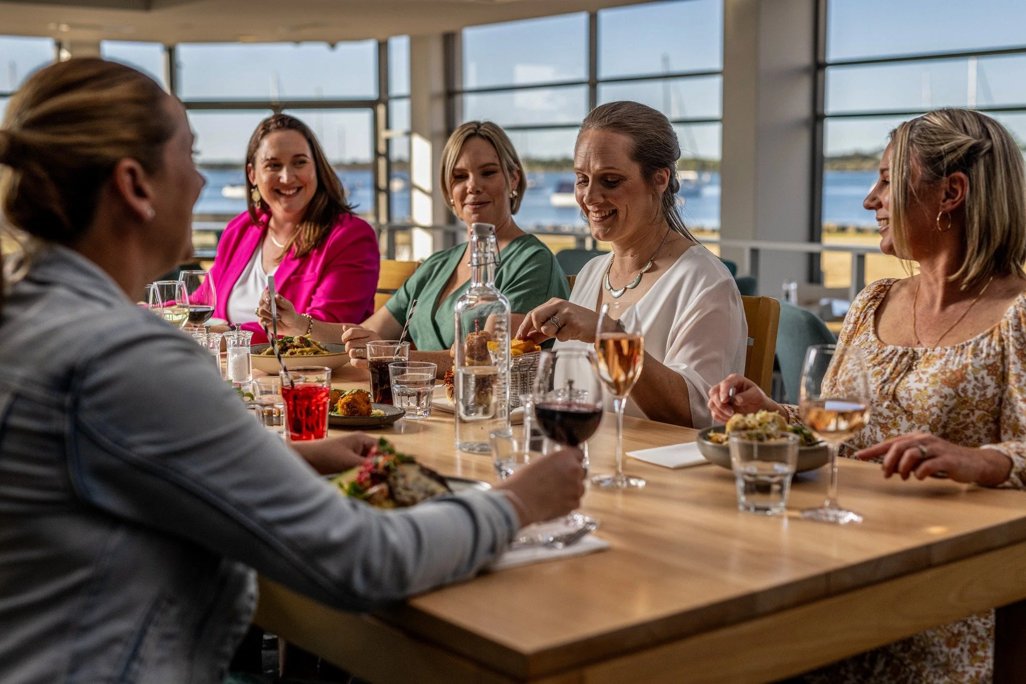 A group of women enjoying a meal together at a restaurant with large windows overlooking a body of water.