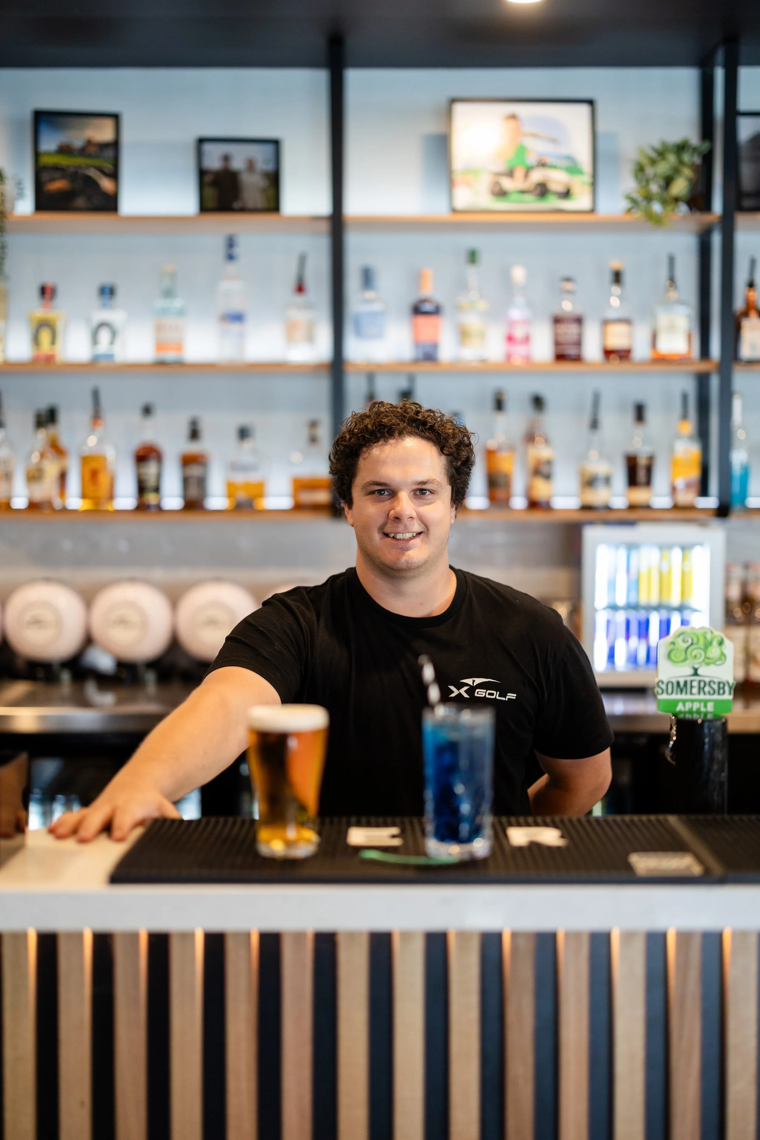 A smiling man with curly hair behind a bar counter with various alcohol bottles in the background, a beer glass in front, and a sign advertising apples.