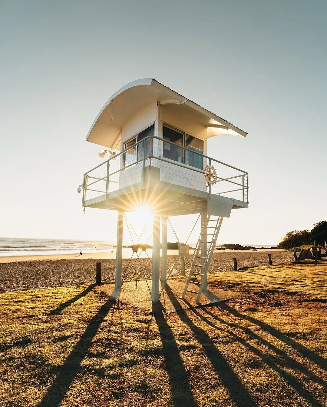A white lifeguard tower on a sandy beach during sunset, with the sun visible behind the tower casting long shadows on the grass and sand.