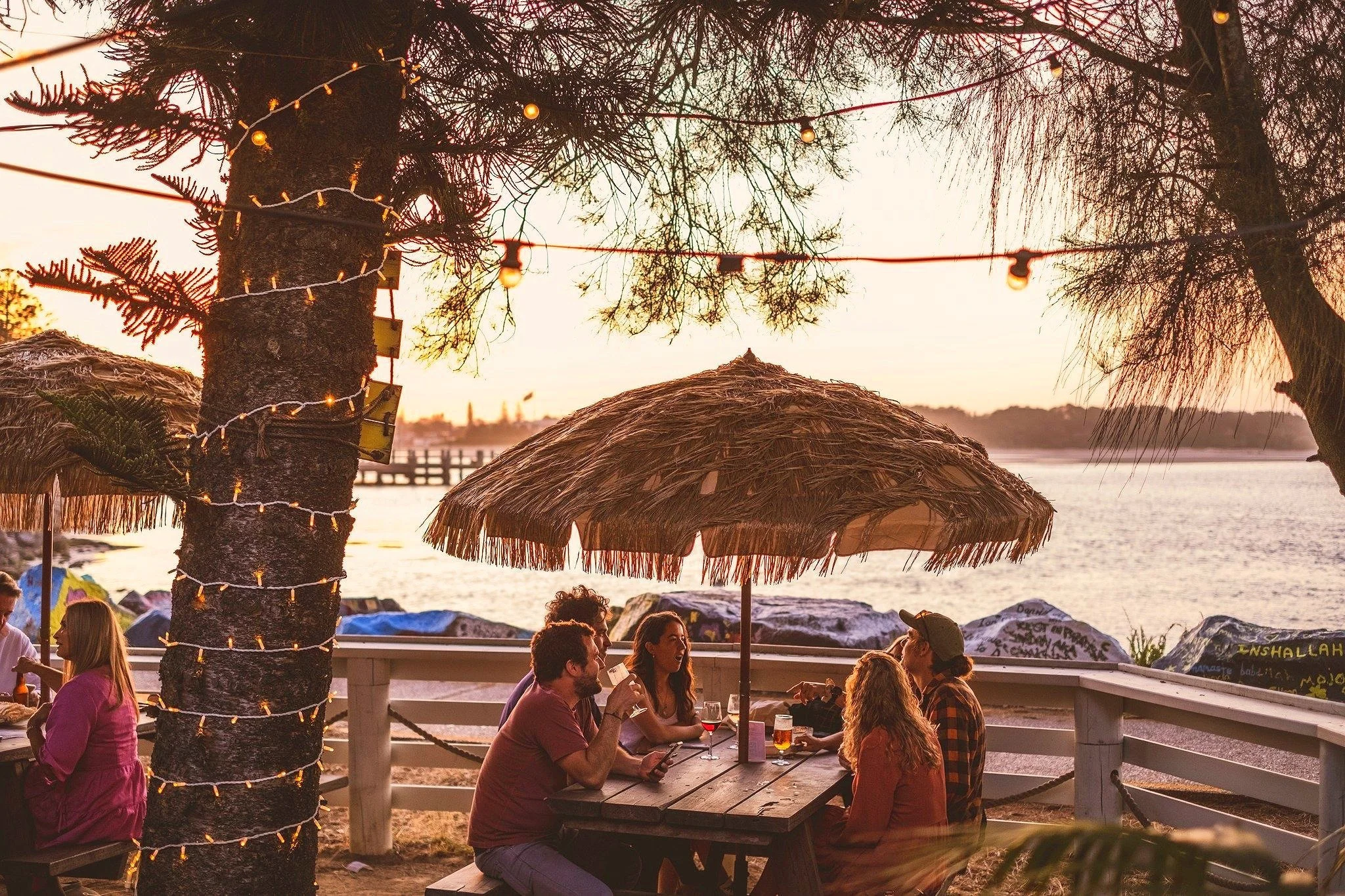 People sitting and talking at outdoor tables under palm umbrellas at sunset, with trees and a body of water in the background, and a tree decorated with string lights.