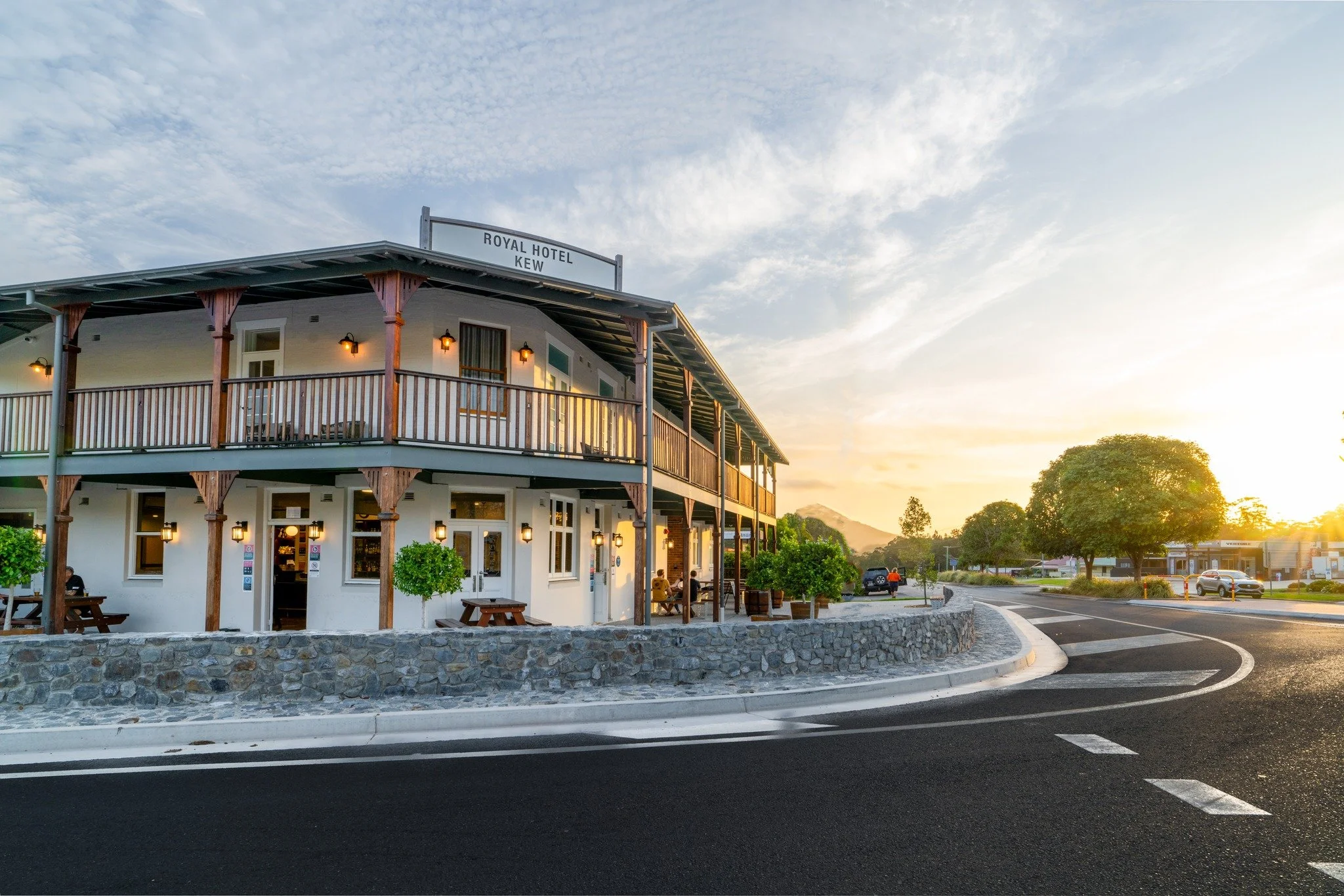 A two-story hotel building with a sign reading "Royal Hotel Kew" on the roof at sunset. The building has a balcony with wooden railing on the upper level and outdoor lighting. There are trees and parking lot with cars and pedestrians nearby, and the sky is partly cloudy.