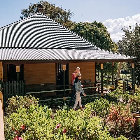 Three people walking past a wooden house with a metal roof in a garden with bushes
