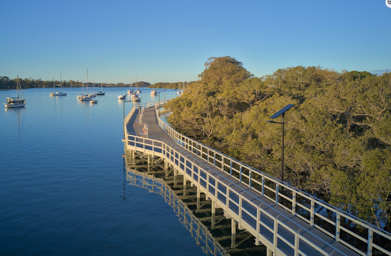 Aerial view of a wooden walkway over water next to a dense green forest, with sailboats anchored nearby under a clear blue sky.