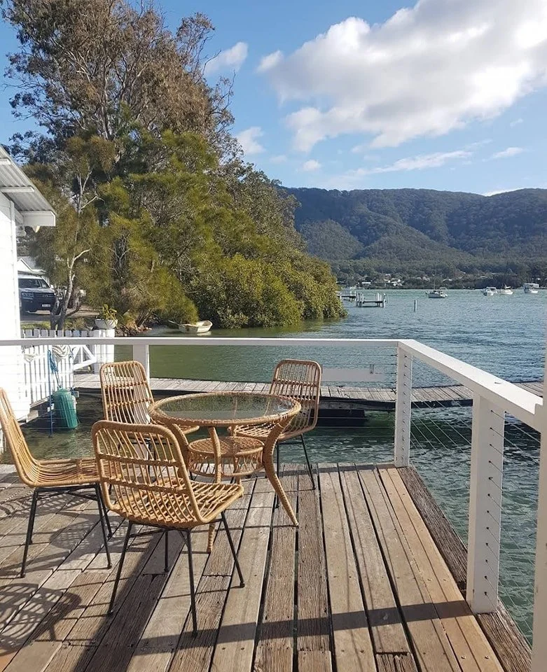 A wooden deck overlooking a lake with a round glass table and four wicker chairs. The lake is calm with several boats and docked boats visible. In the background, trees and hills are seen under a partly cloudy sky.