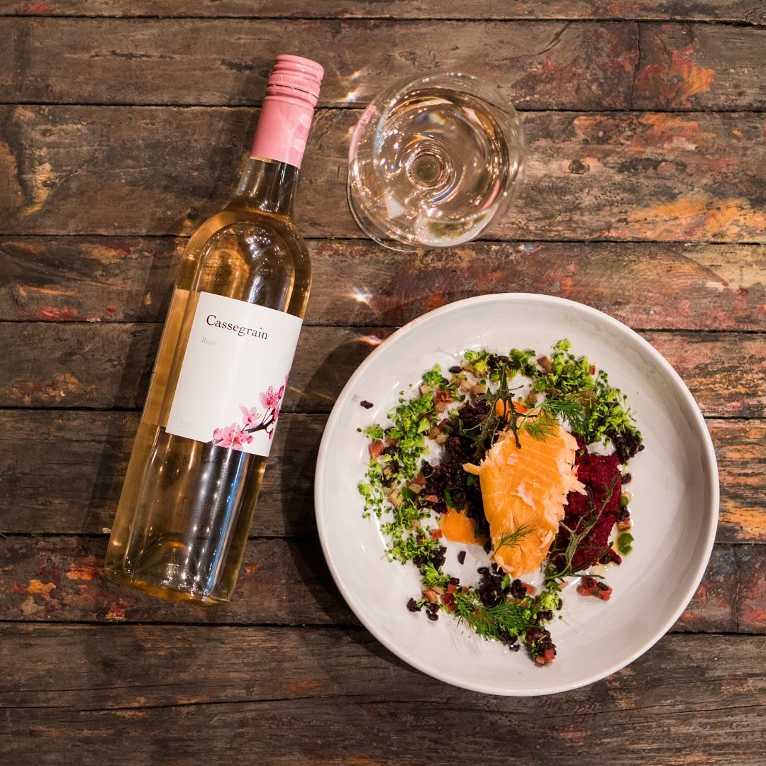 A glass of white wine, a bottle of rosé wine, and a plated salad with salmon, greens, and garnishes on a rustic wooden table.