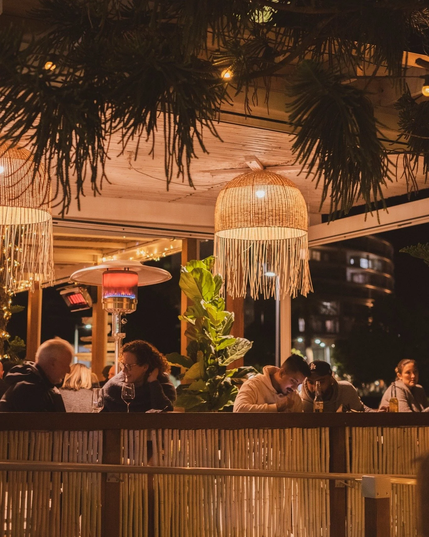 An outdoor restaurant or bar at night with hanging lamps and string lights. Several people are sitting at tables, eating and drinking, with a cityscape visible in the background.